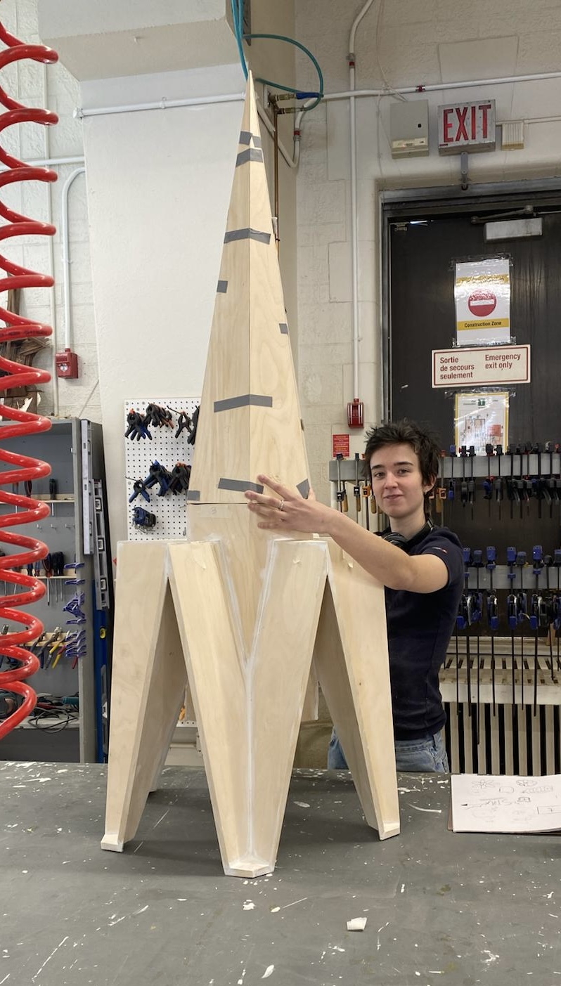 a short haired person in a wood shop stading with a partially constrcted wood sculpture of a small scale gothic church roof