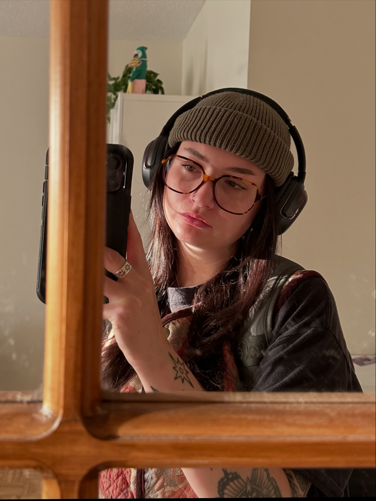 A PERSON WITH LONG DARK HAIR AND A TOQUE AND GLASSES WEARING headphones and holding a phone up to take a selfie and looking into the screen of the phone